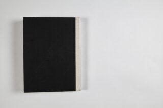 Ein geschlossenes Hardcover-Buch mit schwarzem Einband und hellem Buchrücken liegt auf einer weißen Fläche, positioniert auf der linken Seite des Bildes.