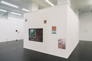 Eine weiße Galeriewand zeigt fünf Fotografien architektonischer Details in einem minimalistischen Ausstellungsraum mit schwarzem Boden und Leuchtstoffröhren an der Decke.