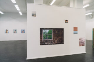 An einer weißen Galeriewand sind verschiedene Fotografien zu sehen, darunter ein großes zentrales Foto eines Fensters mit viel Grün davor, während im Hintergrund weitere Bilder an den angrenzenden Wänden zu sehen sind.