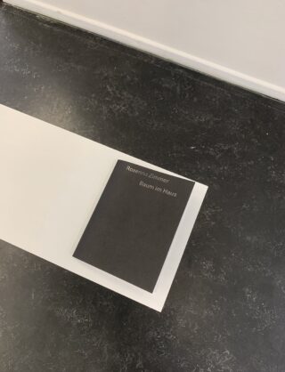 Ein schwarzes Buch mit dem Titel „Roxanna Zimmer Baum im Haus“ liegt auf einer weißen rechteckigen Fläche vor einem dunkel gesprenkelten Boden.