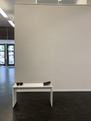 Ein kleiner weißer Tisch mit Kopfhörern und einem geschlossenen Laptop steht vor einer schlichten weißen Wand in einem modernen, minimalistisch eingerichteten Raum.