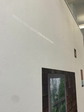 Eine weiße Wand mit dem deutschen Text „Welche Rolle spielen Gefühle bei einer Demenz?“ Die obigen Bilder, darunter ein Gebäudefenster und ein kleines Foto, wahrscheinlich Teil einer Ausstellung.
