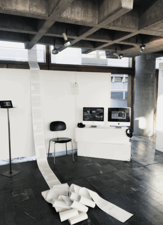 Zwei Computer-Monitore mit Kopfhörern, daneben eine lange Papierbahn, mit Text bedruckt.