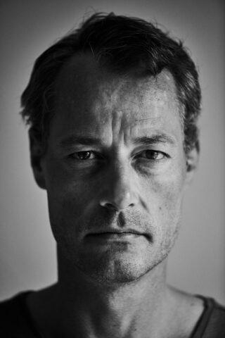 Schwarz-weißes Nahaufnahmeporträt eines Mannes mittleren Alters mit kurzen Haaren, der mit neutralem Gesichtsausdruck direkt in die Kamera blickt.