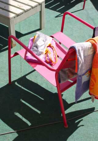 Ein rosa Metallstuhl mit einem Handtuch und einer Tasche darauf steht auf einem grünen Outdoor-Teppich, neben dem ein weiteres Handtuch über der Armlehne hängt. Daneben steht eine Holzbank.