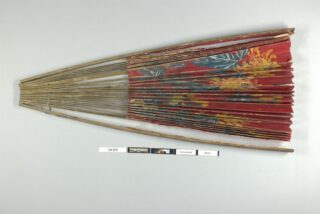 Ein teilweise geöffneter Handfächer mit Bambusrippen und einem rot-blauen Papierblatt mit Blumenmuster, präsentiert auf einer flachen Oberfläche mit Maß- und Identifikationsetiketten darunter.