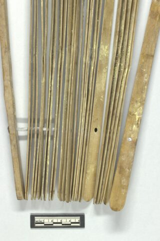 Ein Satz langer, flacher Holzstäbe mit geschnitzten Rillen, vertikal angeordnet, mit einer Maßskala darunter als Größenreferenz.