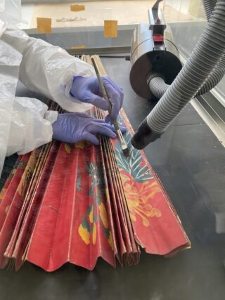 Eine Person mit Handschuhen und Schutzkleidung verwendet ein Werkzeug, um in einer kontrollierten Umgebung einen großen, roten Ventilator mit Blumenmuster neben einem Staubsaugerschlauch zu reparieren oder zu reinigen.