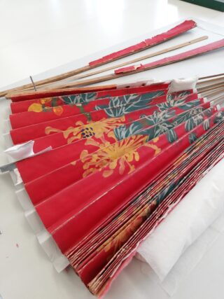 Ein roter Faltfächer aus Papier mit Blumenmuster liegt teilweise geöffnet auf einem Tisch und weist Abnutzungserscheinungen und einige Demontagen auf. In der Nähe liegen Holzrippen und andere Komponenten.