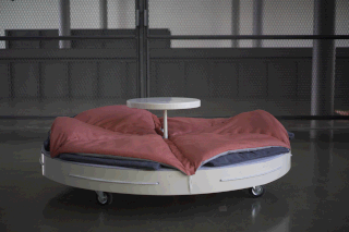 Ein rundes Bett mit roten Decken und blauen Kissen, mit einem kleinen runden Tisch in der Mitte, steht auf Rädern in einer industriellen Innenumgebung.