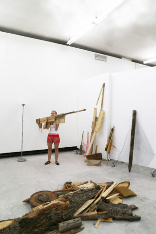 Eine Person steht in einem weißen Raum und hält ein großes Holzobjekt, das einem Gewehr ähnelt, umgeben von verschiedenen Holzgegenständen und Schutt auf dem Boden.