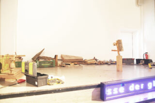 Eine Bühne, auf der Holzbretter, Kisten, Werkzeuge und davor ein LED-Schild verstreut sind; im Hintergrund ist neben einer weißen Wand ein Feuerlöscher zu sehen.