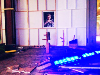 Ein teilweise aufgebauter Raum mit verstreuten Holzbrettern, Werkzeugen, einem leuchtend blauen LED-Schild und einem Poster einer Person an der Rückwand.