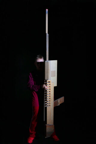 Eine Person in roter Kleidung steht in der Dunkelheit und hält eine hohe, abstrakte, rechteckige Holzstruktur mit verschiedenen Ausschnitten und Vorsprüngen.