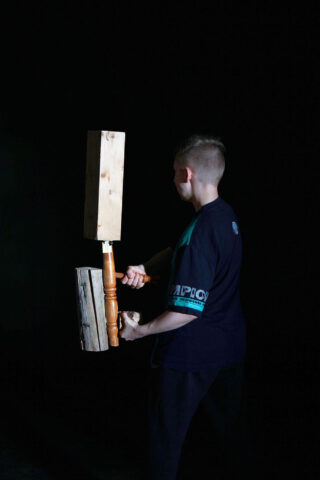 Eine Person hält vor einem schwarzen Hintergrund zwei große Holzhämmer, einen in jeder Hand.