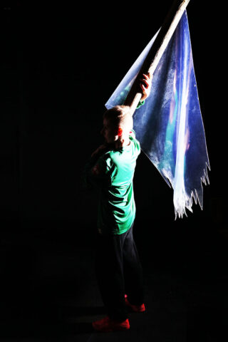 Eine Person in einem grünen Hemd hält eine große, durchscheinende Flagge an einer Stange und steht vor einem dunklen Hintergrund mit dramatischer Beleuchtung.