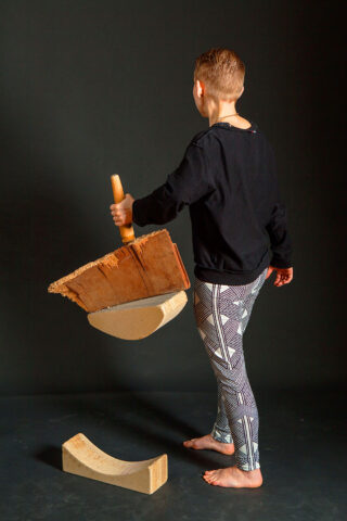 Eine Person in gemusterten Leggings hält ein großes Stück Holz mit einem Griff und steht neben gebogenen und rechteckigen Holzklötzen auf dunklem Hintergrund.