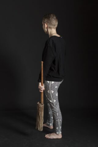 Ein barfüßiges Kind steht mit abgewandtem Gesicht auf einem dunklen Hintergrund, hält einen Holzstab und trägt ein schwarzes Oberteil mit gemusterten Leggings.