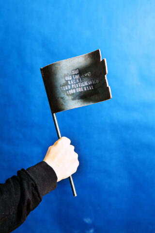 Eine Person hält eine kleine metallische Flagge mit eingraviertem Text vor einem einfarbig blauen Hintergrund.