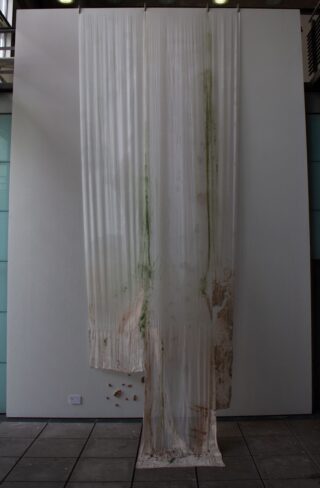 Ein großes, halbtransparentes Laken hängt von der Decke an einer Wand. Es ist mit grünen und braunen Flecken übersät und weist Löcher und ausgefranste Kanten am unteren Rand auf.