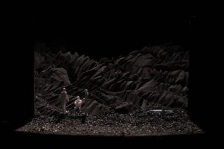 Drei weiße Figuren stehen auf einem dunklen, felsigen Gelände mit gezackten Formationen im Hintergrund und einem kleinen, glänzenden Objekt auf dem Boden rechts.