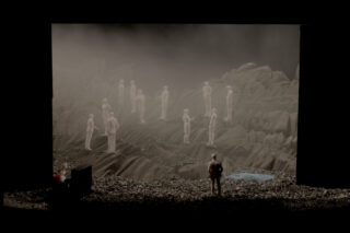 Ein Diorama einer felsigen Landschaft zeigt kleine menschliche Figuren, die einander gegenüberstehen, wobei sich eine Figur in der Nähe eines blauen Teichs und eine andere näher am Vordergrund befindet.