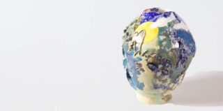 Eine Keramikvase mit einer farbenfrohen, strukturierten Oberfläche mit blauen, grünen, gelben und weißen Mustern auf einem schlichten hellen Hintergrund.