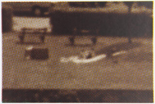 Körniges Schwarzweißbild, das eine Person zeigt, die im Freien auf dem Boden in der Nähe von zwei rechteckigen Objekten, möglicherweise Bänken oder Tischen, liegt.