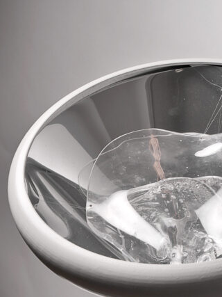 Eine Nahaufnahme eines weißen Keramikspülbeckens mit klarem, zerbrochenem Glas und Wasser darin, vor einem schlichten hellen Hintergrund.