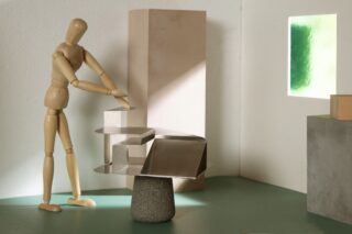 Eine hölzerne Künstlerpuppe steht neben einem modernen, abstrakten Tisch aus Metall und Stein in einem minimalistischen Raum mit sanftem Licht und geometrischem Dekor.