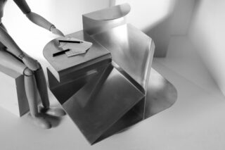 Eine hölzerne Schaufensterpuppe steht in minimalistischem Ambiente neben einem modernen, eckigen Metallschreibtisch, auf dessen Oberfläche zwei Stifte und Blätter Papier liegen.