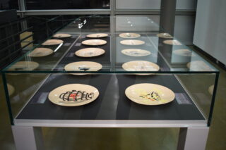 In einer Glasvitrine sind mehrere beige Keramikteller mit verschiedenen abstrakten und farbenfrohen Motiven in Reihen auf einem schwarzen Untergrund angeordnet.