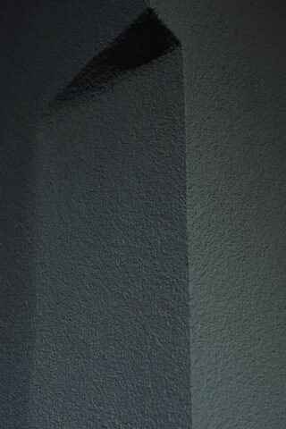 Ein dunkler dreieckiger Schatten wird auf eine strukturierte graue Wand geworfen und bildet scharfe geometrische Linien.