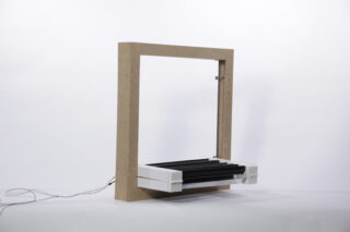Ein rechteckiger Holzrahmen mit weißer Basis hält mehrere schwarze zylindrische Stäbe, von deren linker Seite Drähte ausgehen, vor einem schlichten weißen Hintergrund.