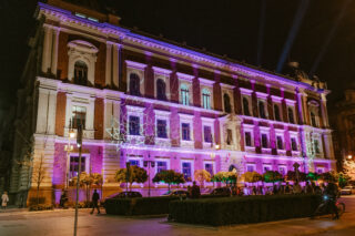 Ein großes, historisches Gebäude, das nachts mit bunten violetten und weißen Lichtern beleuchtet wird, mit Menschen und parkenden Autos davor.