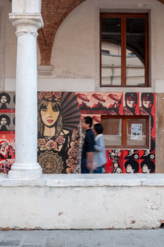 Zwei Menschen gehen an einer Wand mit lebendiger Straßenkunst mit Gesichtern, Blumenmustern und kräftigen Farben vorbei, unter einem gewölbten Gebäude mit einer Säule und einem Fenster mit Holzrahmen.