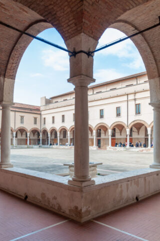 Blick auf einen Innenhof mit gewölbten Gehwegen und Säulen, umgeben von einem zweistöckigen historischen Gebäude unter einem teilweise bewölkten Himmel.