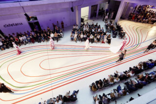 Luftaufnahme einer Modenschau mit Models, die in einem modernen Innenraum entlang bunter Linien auf dem Boden laufen, umgeben von sitzendem Publikum.