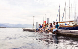 Eine Gruppe von Menschen sitzt auf einem Steg und lässt ihre Füße neben Segelbooten im Wasser baumeln. Im Hintergrund sind Berge zu sehen.