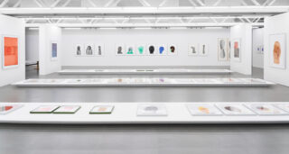 Geräumige Kunstgalerie mit weißen Wänden, in der gerahmte abstrakte Kunstwerke und Porträts ausgestellt sind; weitere Stücke sind auf niedrigen, glasüberdachten Tischen in der Mitte des Raumes angeordnet.