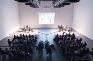 Eine Gruppe von Menschen sitzt in einem modernen Auditorium mit weißen Wänden vor einer Bühne mit Musikern und einem großen Bildschirm, auf dem zwei Blumen zu sehen sind.