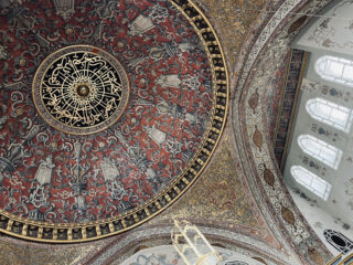 Verzierte, runde Decke mit komplizierten Mustern in Rot, Gold und Schwarz, arabischer Kalligrafie in der Mitte und Bogenfenstern auf der rechten Seite, die natürliches Licht hereinlassen.