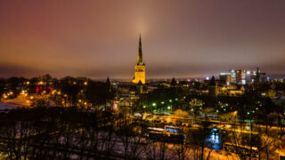 Eine nächtliche Stadtlandschaft mit einem hohen, beleuchteten Kirchturm in der Mitte, umgeben von Gebäuden und Bäumen, und bunten Lichtern, die sich auf den nassen Straßen spiegeln.