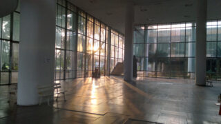 Sonnenlicht strömt durch große Glasfenster in ein geräumiges, modernes Atrium mit weißen Bänken, hohen Säulen und einem Blick auf Bäume und ein Gebäude im Freien.