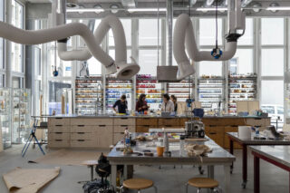 Ein modernes Labor mit großen Fenstern, industriellen Lüftungsschächten und mehreren Menschen, die an zentralen Holztischen arbeiten, umgeben von wissenschaftlichen Geräten und Regalen voller Vorräte.