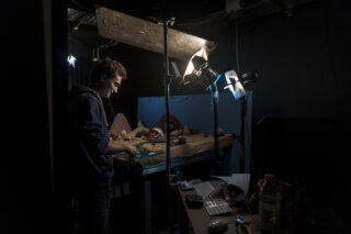 Eine Person arbeitet unter Studiobeleuchtung an einem Miniaturset. In einem schwach beleuchteten Raum sind Kameraausrüstung, Requisiten und eine Computertastatur in der Nähe sichtbar.