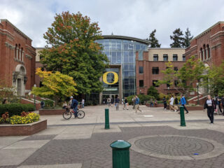 Menschen gehen und fahren mit dem Fahrrad vor einem großen Gebäude mit einem auffälligen gelben „O“-Logo auf dem Campus der University of Oregon.