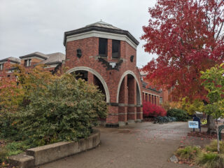 Ein Backsteingebäude mit großen gewölbten Eingängen ist von herbstlich gefärbten Bäumen umgeben. In der Nähe des Eingangs sind ein Schild und Fahrradständer zu sehen.
