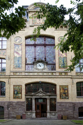 Fassade eines historischen Gebäudes mit großen Buntglasfenstern, dekorativen Kunstwerken, einer zentralen Uhr über dem Eingang und belaubten Ästen, die die Aussicht einrahmen.