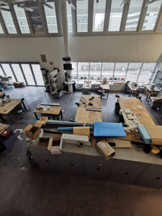 Eine geräumige Werkstatt mit großen Fenstern, mehreren Werkbänken, verstreuten Werkzeugen, Kisten und verschiedenen Materialien, die auf Tischen und Schränken sichtbar sind.